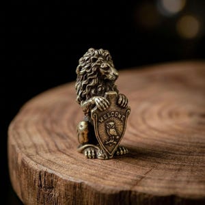May include: A small, antique-style brass figurine of a standing lion with a detailed mane. The lion holds a shield in its front paws, which features an embossed owl and the word WELCOME inscribed in capital letters across the top of the shield.