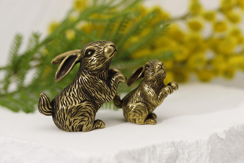 Puede incluir: Dos figuras de conejo de color bronce. Los conejos est&aacute;n representados en una pose juguetona, con las patas delanteras levantadas. Las figuras tienen patrones de pelaje detallados y est&aacute;n colocadas sobre un fondo de flores amarillas y hojas verdes.