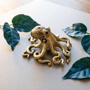 Könnte beinhalten: Eine detaillierte, goldfarbene Oktopus-Figur mit strukturierter Haut und gekräuselten Tentakeln. Der Oktopus ist auf einer hellgrauen Oberfläche zentriert, umgeben von dunkelgrünen Blättern. Die Figur ist ein dekorativer Gegenstand.