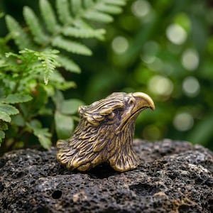 Könnte beinhalten: Eine kleine, goldfarbene Messingfigur in Form eines Adlerkopfes. Die Skulptur zeigt detaillierte, texturierte Federn, die den Hals und Kopf bedecken, sowie einen scharfen, gebogenen Schnabel und ein fokussiertes Auge im Profil des Vogels.