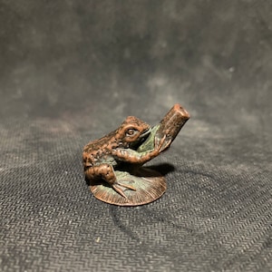 May include: A small bronze frog figurine sitting on a round base, holding a pipe in its mouth.