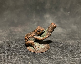 May include: A small bronze frog figurine sitting on a round base, holding a pipe in its mouth.