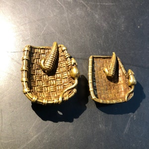 Pode incluir: Duas peças decorativas de metal dourado em forma de cestas com um design de espiga de milho. As cestas têm um padrão tecido e são detalhadas com pequenas saliências em relevo.