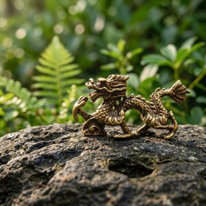 Puede incluir: Una peque&ntilde;a figura de drag&oacute;n de metal color lat&oacute;n con detalles intrincados de escamas, un cuerpo curvado y una cola levantada. La criatura m&iacute;tica se muestra en una pose din&aacute;mica con la cabeza girada, destacando sus texturas y acabado.