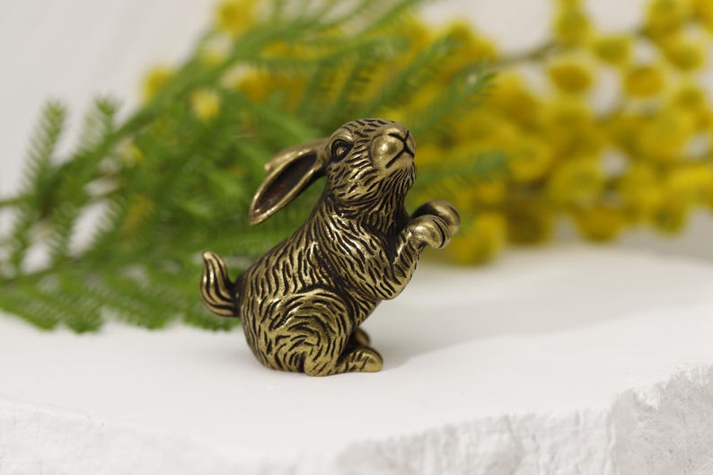 Puede incluir: Una figura de conejo de color bronce en posici&oacute;n sentada, con las patas delanteras levantadas. El conejo tiene una textura de pelaje detallada y orejas largas. El fondo presenta follaje verde y flores amarillas.