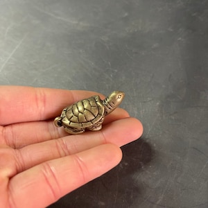 Pode incluir: Pequena figura de tartaruga em latão de estilo antigo. A tartaruga tem um padrão de carapaça detalhado e uma cabeça pequena com olhos visíveis. A figura é de uma cor metálica quente e parece ser feita de metal sólido.