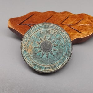 Puede incluir: Una moneda redonda de cobre con un diseño de sol radiante y caracteres chinos alrededor del borde. La moneda tiene una pátina verde.
