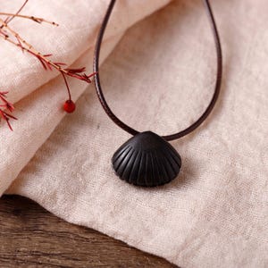 May include: A black seashell pendant necklace with a brown cord. The pendant has a detailed, ridged design. The necklace is displayed on a beige fabric surface.