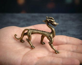 May include: A small, antique-style brass figurine of a dragon with a slender, elongated body, textured scales, four legs with clawed feet, and a curved tail. The metallic surface features a dark, weathered patina with golden highlights.