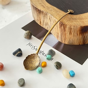 May include: A gold-coloured spoon with an ornate design on the bowl. The spoon is sitting on a white surface with small stones and a piece of paper with the text "YVES SAINT LAURENT" visible.