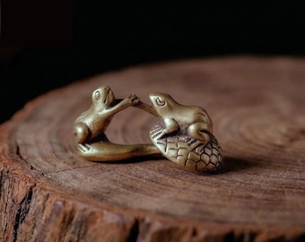 Könnte beinhalten: Eine kleine Messingfigur, die zwei interagierende Frösche darstellt. Ein Frosch sitzt auf einer strukturierten, abgerundeten Basis, während der andere Frosch nach ihm greift. Das Metall hat einen warmen Goldton mit glatter Oberfläche.