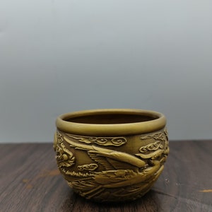 Puede incluir: Una pequeña taza de metal de color dorado con intrincados diseños en relieve de pájaros y nubes. La taza está sobre una superficie de madera.
