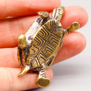 May include: A small, detailed brass turtle figurine. The turtle is lying on its back, with its legs and head extended. The shell features a geometric pattern. The figurine has a vintage, antique appearance.