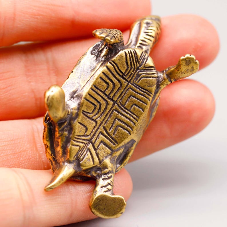May include: A small, detailed brass turtle figurine. The turtle is lying on its back, with its legs and head extended. The shell features a geometric pattern. The figurine has a vintage, antique appearance.
