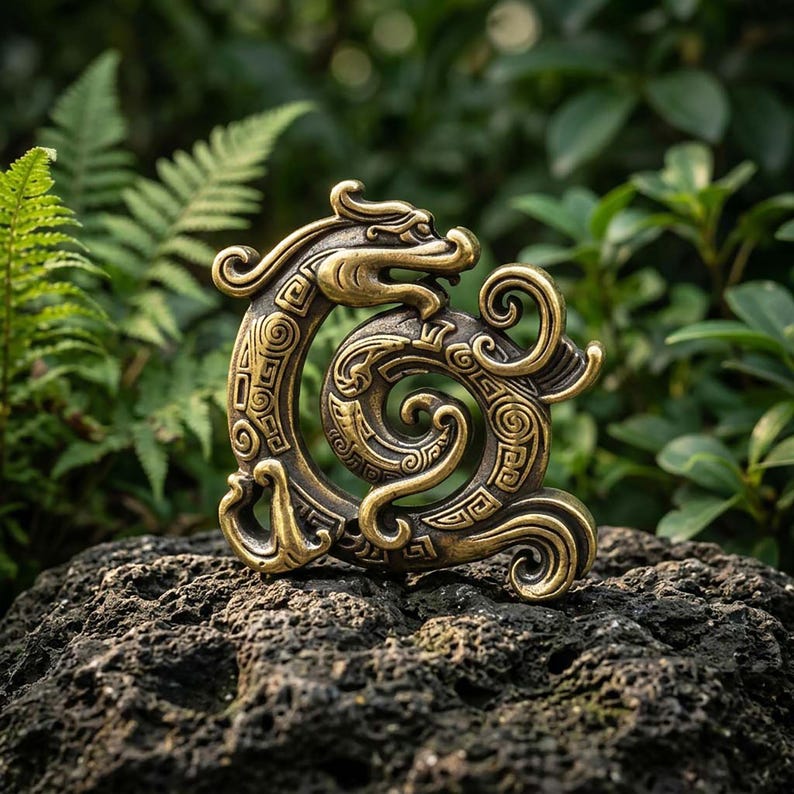 Puede incluir: Una figura de drag&oacute;n de metal en tono dorado con forma circular y enroscada. La superficie presenta intrincados patrones geom&eacute;tricos en relieve y l&iacute;neas onduladas por todo el cuerpo, con una cabeza de drag&oacute;n prominente en la parte superior.