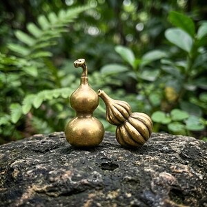 May include: Two small brass gourd-shaped ornaments. One gourd is smooth with a curved stem, while the other features vertical ridges and a tapered neck. Both items have a metallic gold finish and are positioned upright on a textured surface.