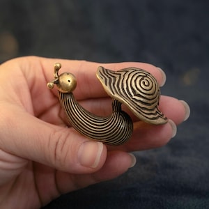 May include: A small brass figurine shaped like a snail with a smooth, rounded head featuring two antennae and a shell decorated with a detailed, raised spiral pattern. The body of the snail is textured with fine, horizontal lines throughout.