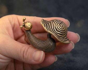 May include: A small brass figurine shaped like a snail with a smooth, rounded head featuring two antennae and a shell decorated with a detailed, raised spiral pattern. The body of the snail is textured with fine, horizontal lines throughout.