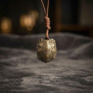 Puede incluir: Un colgante de metal en tono latón con forma de tortuga y un patrón hexagonal detallado en el caparazón. El colgante cuelga de un cordón marrón que incluye un nudo ajustable en la parte superior, creando una pieza de joyería funcional.
