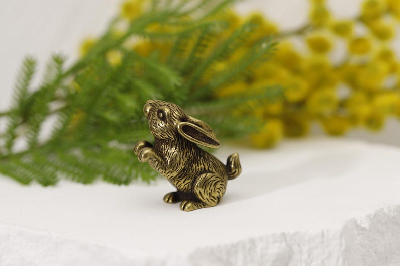 Puede incluir: Una detallada figura de conejo de bronce, posando de pie con las patas delanteras levantadas. El conejito tiene pelaje texturizado y orejas largas. El fondo presenta follaje verde y flores amarillas. La figura est&aacute; sobre una superficie blanca.