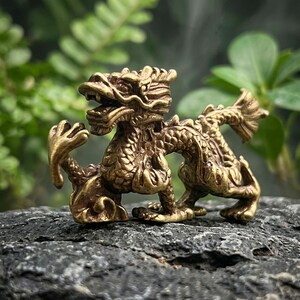 Puede incluir: Una peque&ntilde;a figura de drag&oacute;n de metal dorado con texturas de escamas intrincadas, una cabeza detallada con la boca abierta y una cola curva. La criatura se muestra en una pose din&aacute;mica con una garra delantera levantada y extendida.
