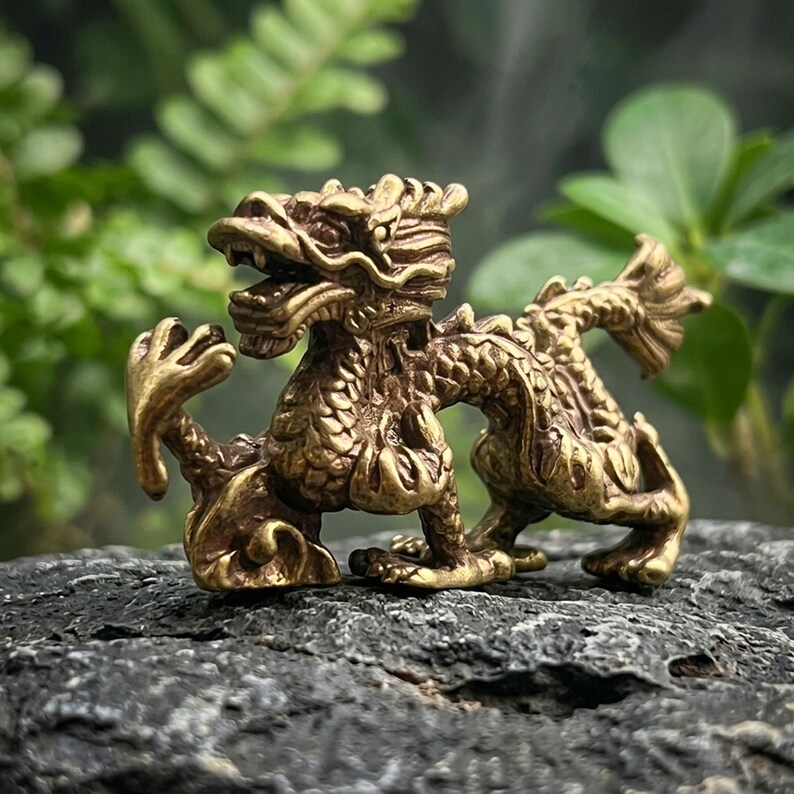 Puede incluir: Una peque&ntilde;a figura de drag&oacute;n de metal dorado con texturas de escamas intrincadas, una cabeza detallada con la boca abierta y una cola curva. La criatura se muestra en una pose din&aacute;mica con una garra delantera levantada y extendida.