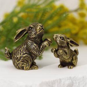 Puede incluir: Dos figuras de conejo de bronce. Un conejo est&aacute; erguido, con las patas levantadas, y el otro es m&aacute;s peque&ntilde;o y lo mira. Los conejos tienen texturas de pelaje detalladas, sobre un fondo de follaje amarillo y verde.