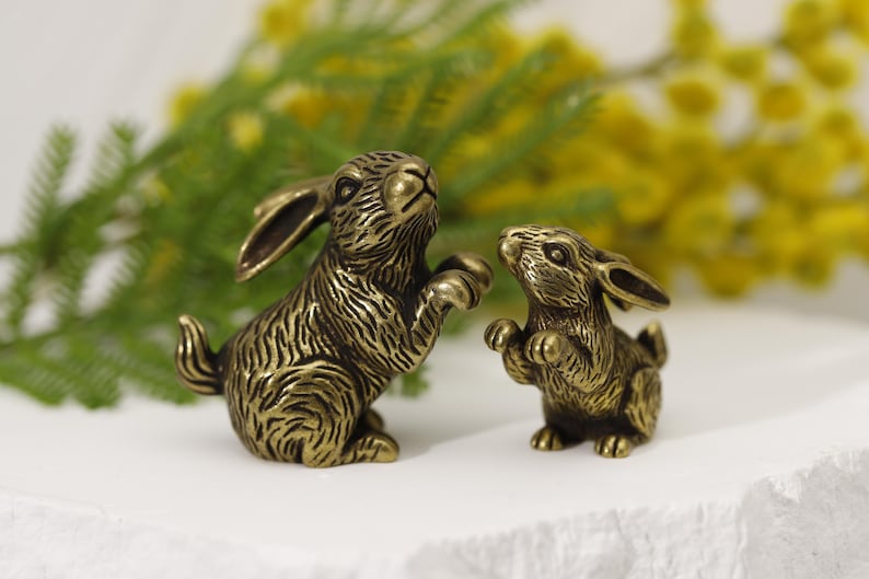 Puede incluir: Dos figuras de conejo de bronce. Un conejo est&aacute; erguido, con las patas levantadas, y el otro es m&aacute;s peque&ntilde;o y lo mira. Los conejos tienen texturas de pelaje detalladas, sobre un fondo de follaje amarillo y verde.