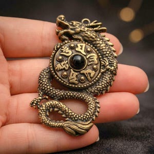 May include: A brass-toned metal pendant shaped like a coiled dragon surrounding a circular medallion. The medallion features embossed Tibetan script characters arranged around a central, smooth, polished black stone bead set in the middle.