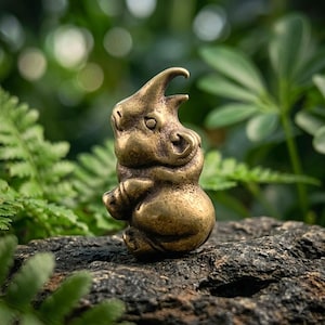 May include: A small, solid brass figurine of a rhinoceros with a smooth, metallic finish. The rhinoceros is depicted in a seated, upright posture with its head turned slightly to the side, showcasing detailed facial features and a prominent horn.