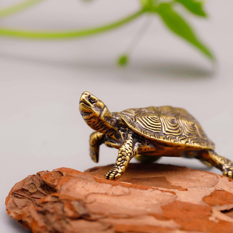 May include: A detailed bronze turtle figurine with intricate shell patterns. The turtle is posed on a piece of brown bark, with a blurred green background. The figurine is a decorative item.