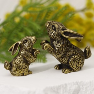 Puede incluir: Dos figuras de conejos de color bronce. Un conejo est&aacute; de pie, el otro es m&aacute;s peque&ntilde;o y en una pose similar. El fondo presenta follaje verde y flores amarillas, sobre una superficie blanca.