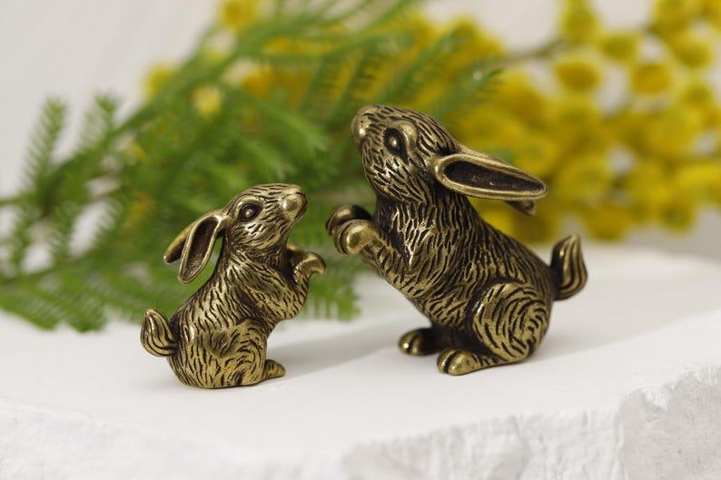 Puede incluir: Dos figuras de conejos de color bronce. Un conejo est&aacute; de pie, el otro es m&aacute;s peque&ntilde;o y en una pose similar. El fondo presenta follaje verde y flores amarillas, sobre una superficie blanca.