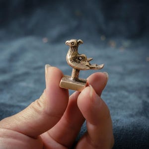 以下が含まれることがあります： 翼が様式化され、頭を横に向けた鳥の形をした、小さなブロンズ色の金属製フィギュア。鳥は長方形の台座に取り付けられており、胴体には線が刻まれ、翼には円形の切り抜き模様が施されている。