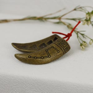 May include: A pair of gold-toned metal earrings shaped like horns with a constellation design and a Greek key pattern. The earrings are connected by a red string.