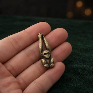Könnte beinhalten: Eine kleine Messingfigur eines Affen mit über den Kopf erhobenen Armen. Der Affe weist detaillierte Fellstrukturen, ein glattes Gesicht und ein metallisch goldenes Finish auf. Die Figur ist kompakt und mit einer Schlaufe durch die Arme gestaltet.