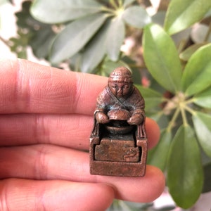 Puede incluir: Una pequeña estatua de bronce de una persona sentada en un trono sosteniendo un cuenco. La estatua es detallada y tiene un acabado desgastado.