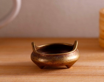 Könnte beinhalten: Ein kleiner Räuchergefäß aus Messing im antiken Stil. Es hat eine abgerundete Schalenform, drei kleine Füße und zwei gebogene Griffe. Das Messing hat eine warme, gealterte Patina, die darauf hindeutet, dass es aus der Zeit stammt. Der Brenner steht auf einer hellen Holzoberfläche.