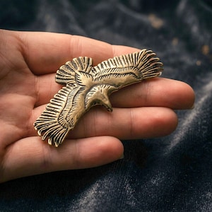 Puede incluir: Un broche de águila en metal dorado con detallados patrones de plumas grabados en las alas y el cuerpo. El ave se muestra en posición de vuelo con las alas extendidas, exhibiendo un acabado texturizado y un perfil de pico definido.