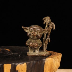 Puede incluir: Una pequeña figura de bronce de un cerdo sosteniendo un objeto redondo, de pie junto a un tallo de bambú con hojas. El cerdo tiene una cara sonriente y está representado en un estilo de dibujos animados.