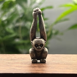 May include: A silver figurine of a gibbon monkey with its arms raised above its head. The monkey is sitting on a wooden surface.