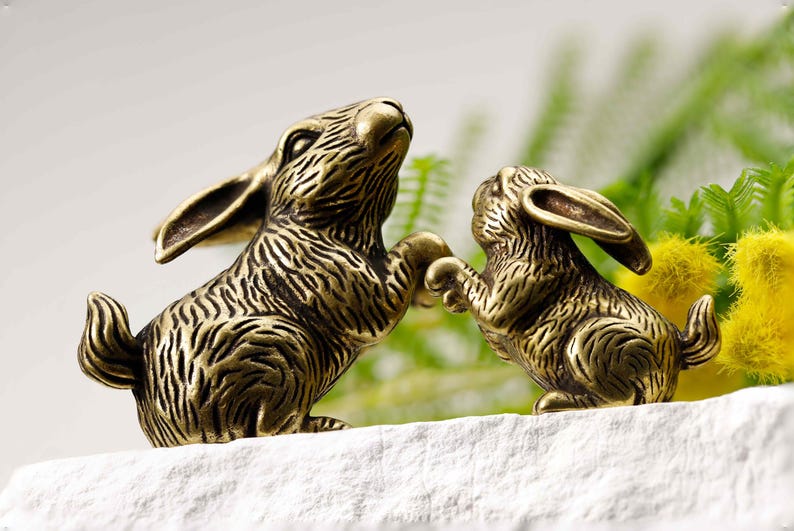 Puede incluir: Dos figuras de conejos de bronce, una mirando hacia arriba y la otra extendiendo la mano. Los conejos tienen pelaje texturizado y est&aacute;n colocados sobre una superficie blanca, con un fondo verde y amarillo borroso.