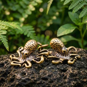 May include: Two metallic octopus figurines with a bronze finish and textured, spotted heads. Each octopus features multiple curved, detailed tentacles extending outward, resting on a dark, porous, uneven rock surface with green foliage.