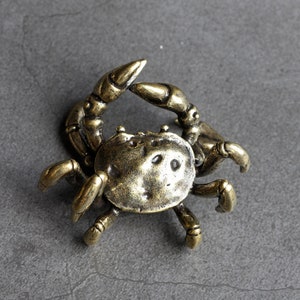 Puede incluir: Una pequeña figura de cangrejo de latón detallada. El cangrejo está en posición con sus garras levantadas y sus patas extendidas.