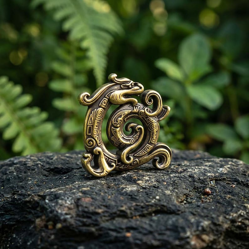 Puede incluir: Un adorno de metal color lat&oacute;n con forma de drag&oacute;n enroscado con intrincados patrones geom&eacute;tricos en relieve y l&iacute;neas onduladas. El objeto presenta una cabeza estilizada en la parte superior y un cuerpo serpentino curvo en espiral.