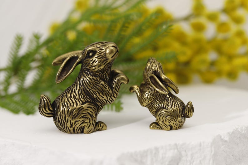 Puede incluir: Dos figuras de conejo de bronce. Un conejo est&aacute; erguido, el otro en una pose similar. Los conejos tienen una textura de pelaje detallada y est&aacute;n colocados sobre un fondo de flores amarillas y follaje verde. Las figuras est&aacute;n sobre una superficie blanca.