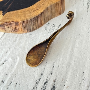 May include: A brass spoon with a bird-shaped handle. The spoon has a long, curved stem and a deep, oval bowl. The brass has a warm, aged patina. The spoon is set against a white surface with a wooden block in the background.