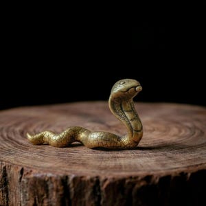 Op de afbeelding: Een klein koperen slangenbeeldje met een textuur van gebarsten patronen. De slang is afgebeeld in een rechtopstaande, alerte houding met de kop omhoog en de nek gespreid, met een metallic gouden afwerking tegen een donkere achtergrond.