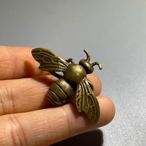 Puede incluir: Un broche de abeja de bronce con alas detalladas y un cuerpo texturizado. La abeja está en posición de vuelo con las alas extendidas.
