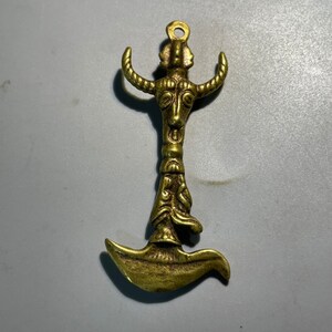 May include: A gold-colored metal pendant featuring a stylized bull head with horns and a decorative design. The pendant is shaped like a crescent moon with a loop at the top for hanging.